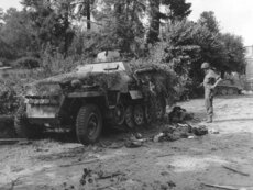 Un GI armé d'un M3 " Grease gun " regarde le cadavre d'un soldat allemand derrière un semi-chenillé allemand SdKfz 251 Ausf. D. camouflé avec des branchages.